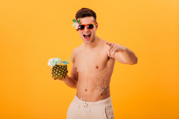 Alegre hombre desnudo en pantalones cortos y gafas de sol inusuales con cóctel mientras apunta y mira a la cámara sobre amarillo