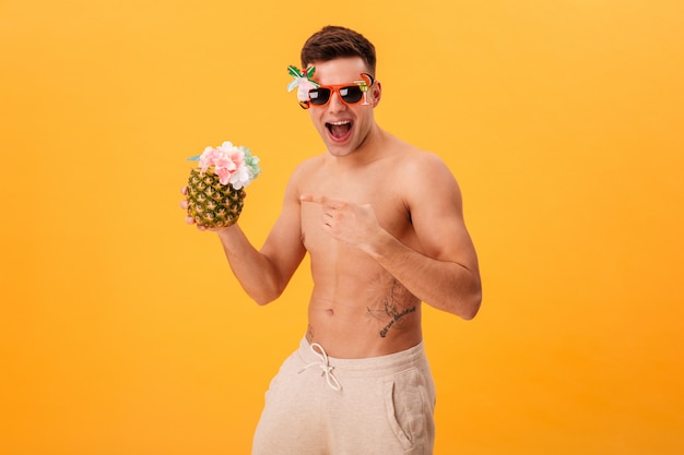 Alegre hombre desnudo en pantalones cortos y gafas de sol inusuales con cóctel mientras apunta sobre él y mira la cámara sobre amarillo
