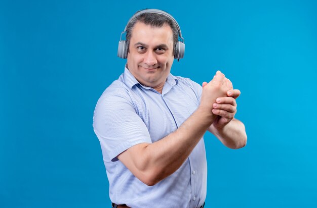 Alegre hombre de mediana edad en camisa azul a rayas con audífonos sosteniendo sus manos juntas mientras está en un espacio azul