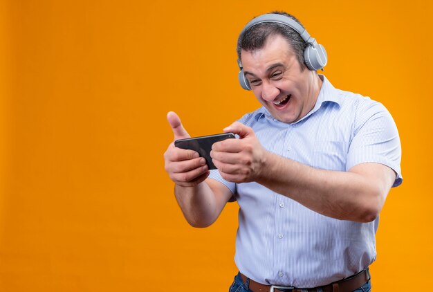 Alegre hombre de mediana edad vestido con camisa azul de rayas verticales jugando en el teléfono móvil con auriculares mientras está de pie