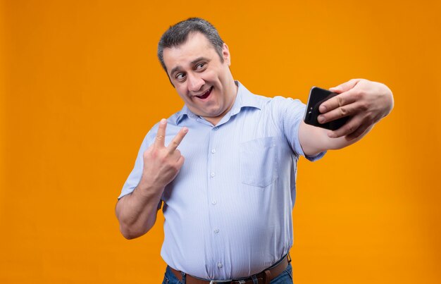 Alegre hombre de mediana edad vestido con camisa azul a rayas verticales riendo y tomando selfie en smartphone