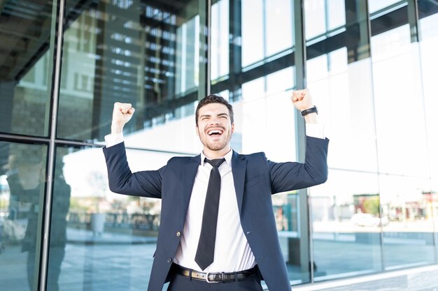 Alegre hombre de negocios hispano levantando los brazos y celebrando la victoria fuera del edificio de oficinas