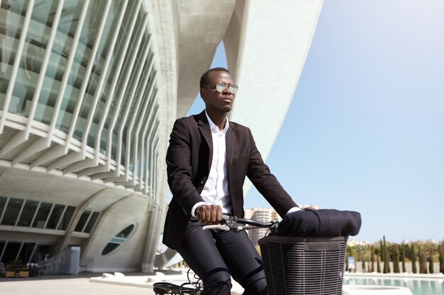 Alegre hombre de negocios masculino europeo negro elegante que usa la bicicleta para llegar a la oficina mientras su automóvil está roto, en bicicleta en un entorno urbano, pasando por edificios modernos y una fuente en un día soleado de verano