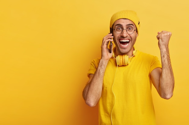 Alegre hombre optimista con una gran sonrisa, levanta el puño cerrado en señal de triunfo, llama a través de un teléfono inteligente, disfruta de la conversación