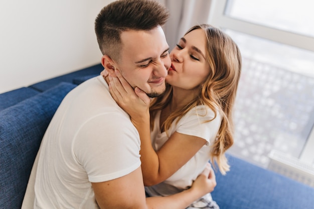 Alegre hombre de pelo oscuro en traje blanco pasando la mañana con su esposa. Adorable chica rubia besando a novio en fin de semana.