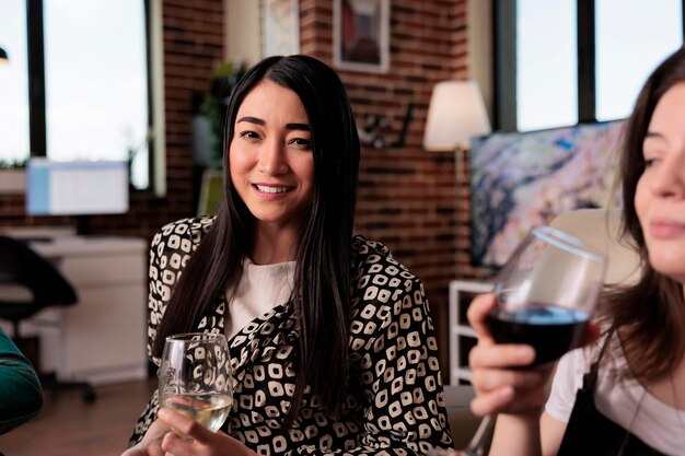 Alegre joven adulto disfrutando de una copa de vino en un evento de celebración de la amistad mientras se sienta en la sala de estar. Mujer asiática feliz disfrutando de la fiesta del vino con los mejores amigos mientras sonríe a la cámara.