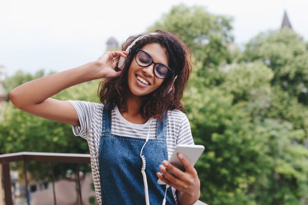 Alegre joven africana en camisa a rayas de pie en el árbol y escuchar la canción con una sonrisa y los ojos cerrados