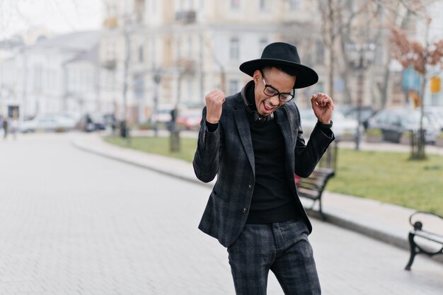 Alegre joven africano feliz posando en el parque por la mañana. Retrato al aire libre de chico delgado y elegante en traje a cuadros que expresa emociones positivas.