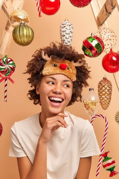 Alegre joven afroamericana mantiene la mano debajo de la barbilla, sonríe con dientes, tiene un ambiente festivo, va a colgar adornos navideños en el abeto, disfruta de un ambiente festivo acogedor, usa camiseta y antifaz