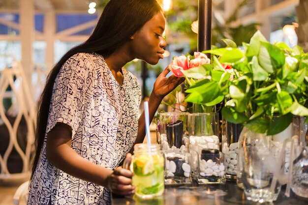 Alegre joven afroamericana en vestido de verano en el café huele flores blancas en florero.