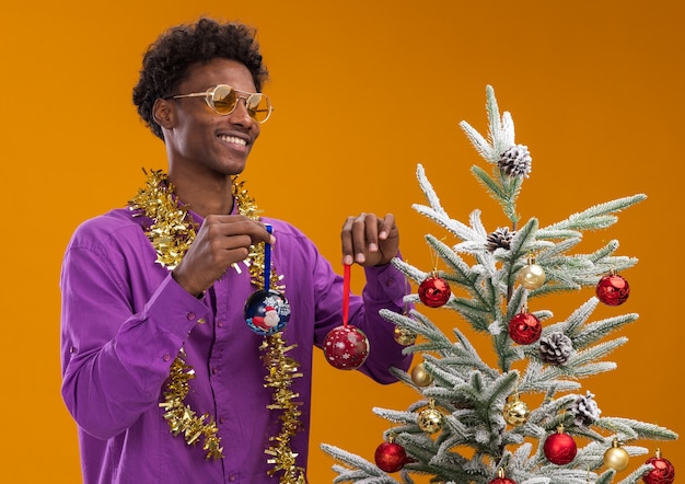 Alegre joven afroamericano con gafas con guirnalda de oropel alrededor del cuello de pie cerca del árbol de Navidad decorado sobre fondo naranja