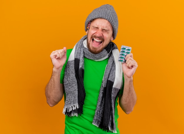 Alegre joven apuesto enfermo eslavo con gorro y bufanda de invierno, sosteniendo un paquete de cápsulas, haciendo un gesto de sí con los ojos cerrados