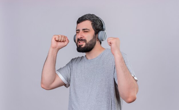 Alegre joven apuesto hombre caucásico con auriculares escuchando música con los ojos cerrados y los puños levantados aislados en blanco