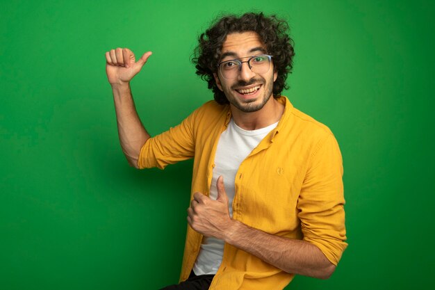 Alegre joven apuesto hombre caucásico con gafas mostrando los pulgares para arriba aislado en la pared verde con espacio de copia