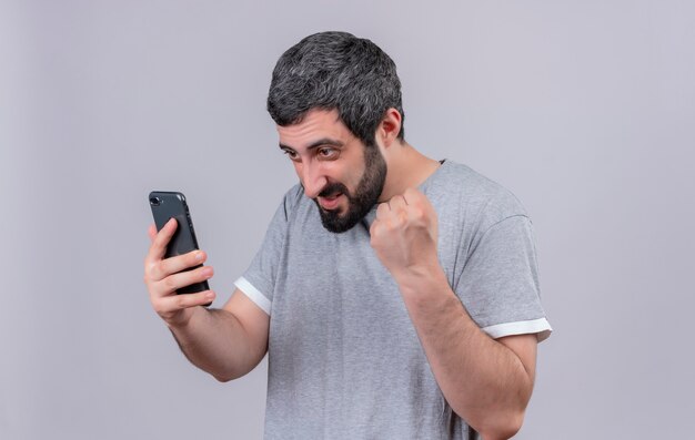 Alegre joven apuesto hombre caucásico sosteniendo y mirando el teléfono móvil y levantando el puño aislado en blanco