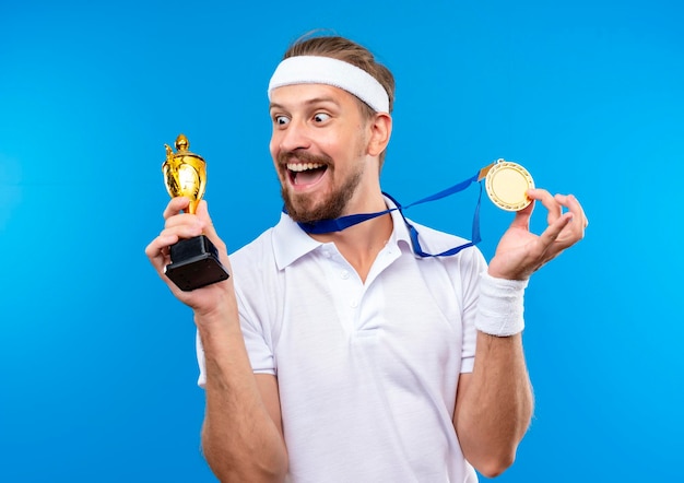 Alegre joven apuesto hombre deportivo con diadema y muñequeras y medalla alrededor del cuello sosteniendo la medalla y la copa ganadora y mirando la copa aislada en la pared azul