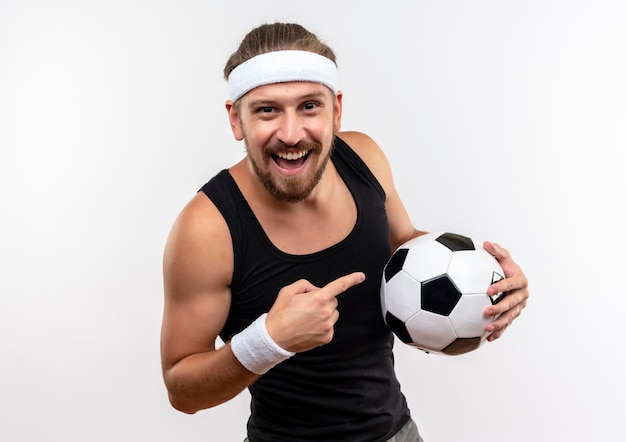 Foto gratuita alegre joven apuesto hombre deportivo con diadema y muñequeras sosteniendo y apuntando a un balón de fútbol aislado en la pared blanca