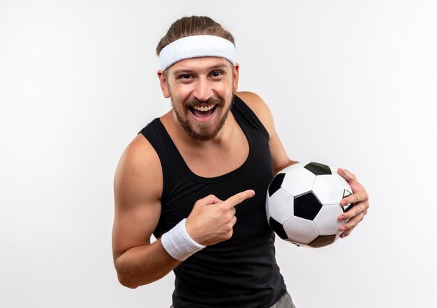 Alegre joven apuesto hombre deportivo con diadema y muñequeras sosteniendo y apuntando a un balón de fútbol aislado en la pared blanca