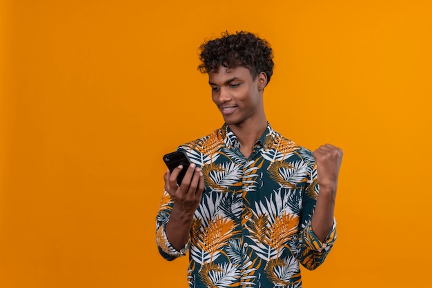 Alegre joven apuesto hombre de piel oscura con cabello rizado en hojas de camisa estampada mirando smartphone con puño apretado