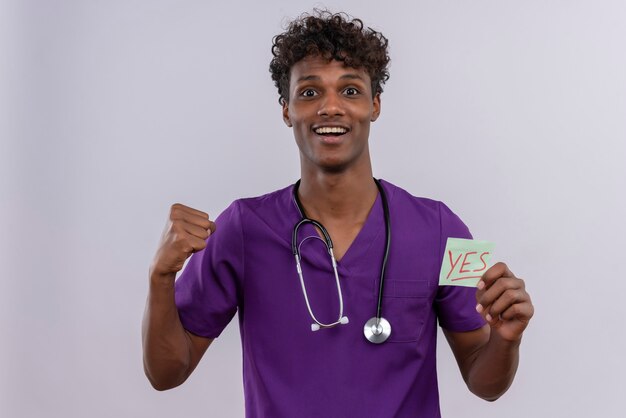 Un alegre joven apuesto médico de piel oscura con cabello rizado vistiendo uniforme violeta con estetoscopio mostrando una tarjeta de papel con la palabra sí