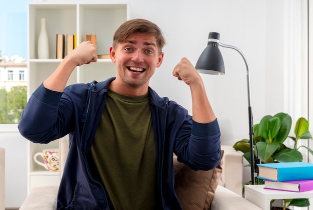 Alegre joven apuesto rubio se sienta en un sillón manteniendo los puños mirando a la cámara dentro de la sala de estar