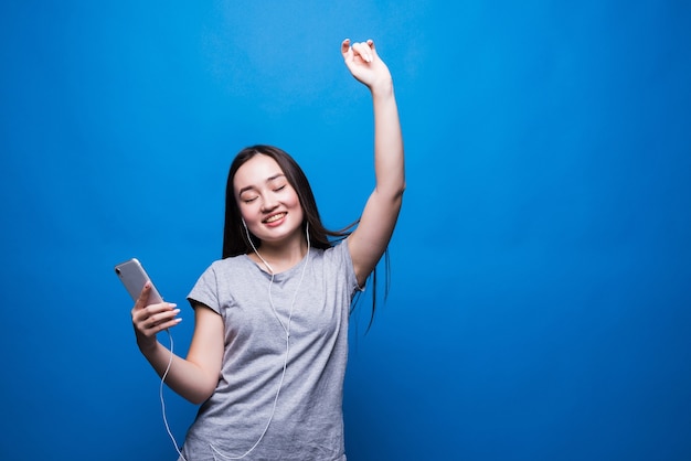 Alegre joven asiática en auriculares escuchando música y bailando aislado sobre la pared azul