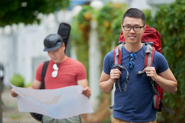 Alegre joven asiática posando con mochila y amigo mirando el mapa