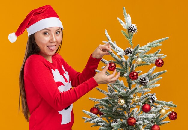 Alegre joven asiática con sombrero de navidad con suéter decorar el árbol de navidad mostrando la lengua aislada en la pared naranja