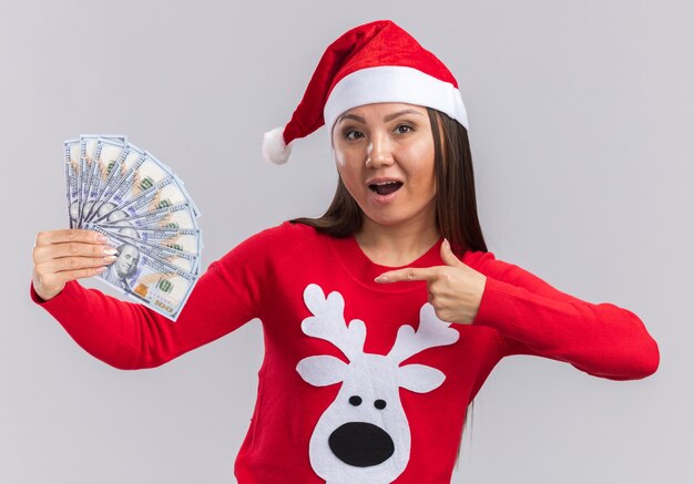 Alegre joven asiática vistiendo gorro de navidad con suéter sosteniendo y puntos en efectivo aislado sobre fondo blanco.