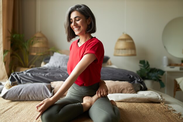 Alegre joven atlética sentada descalza sobre la alfombra, manteniendo las piernas cruzadas, haciendo un giro espinal sentado durante la clase de yoga, disfrutando del ejercicio de estiramiento, respirando profundamente, cerrando los ojos