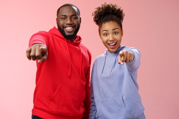 Alegre joven atractivo feliz pareja afroamericana de vacaciones disfrutando de un interesante recorrido de pie impresionado asombrado fondo rosa apuntando con los dedos índice cámara impresionante objeto curioso