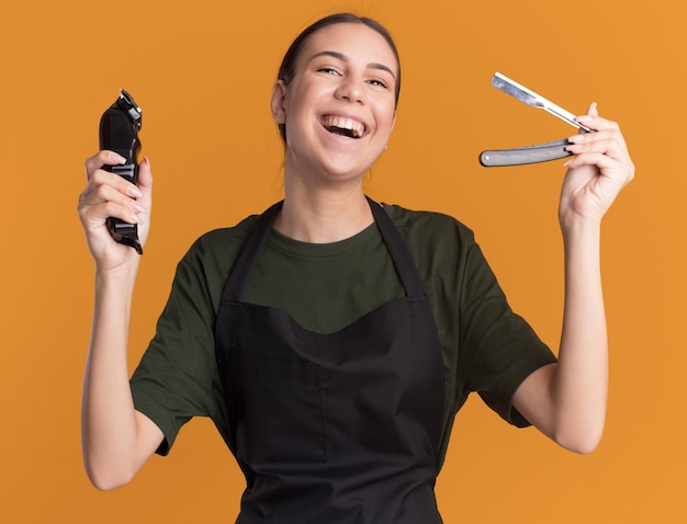 Alegre joven barbero morena en uniforme sosteniendo cortapelos y navaja de afeitar recta