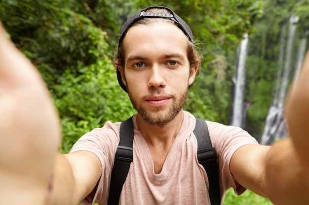 Alegre joven barbudo hipster con gorra de béisbol tomando selfie en sus exóticas vacaciones