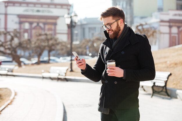 Alegre joven barbudo usando teléfono celular y tomando café en la ciudad