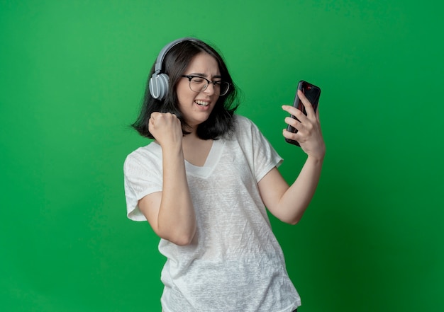 Alegre joven bastante caucásica con gafas y auriculares sosteniendo el teléfono móvil apretando el puño con los ojos cerrados aislado sobre fondo verde con espacio de copia