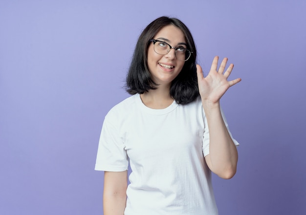 Alegre joven bastante caucásica con gafas mirando hacia arriba y mostrando cinco con la mano aislada sobre fondo púrpura con espacio de copia