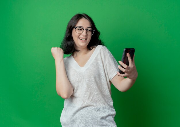 Alegre joven bastante caucásica con gafas sosteniendo y mirando el teléfono móvil y apretando el puño