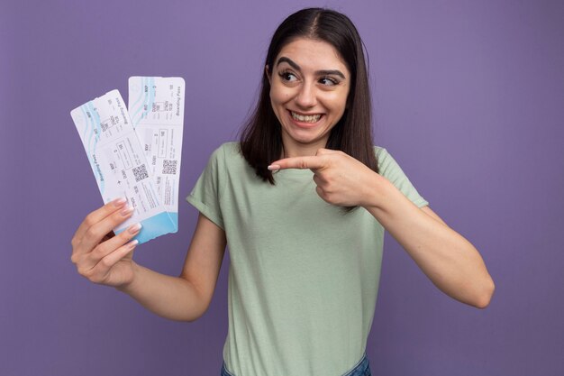 Alegre joven bastante caucásica sosteniendo y apuntando a los billetes de avión mirando al lado aislado en la pared púrpura