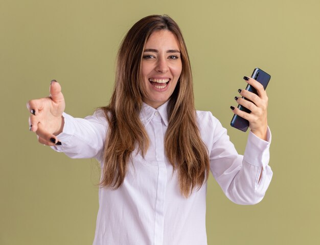 Alegre joven bastante caucásica sosteniendo el teléfono y estirando la mano aislada en la pared verde oliva con espacio de copia