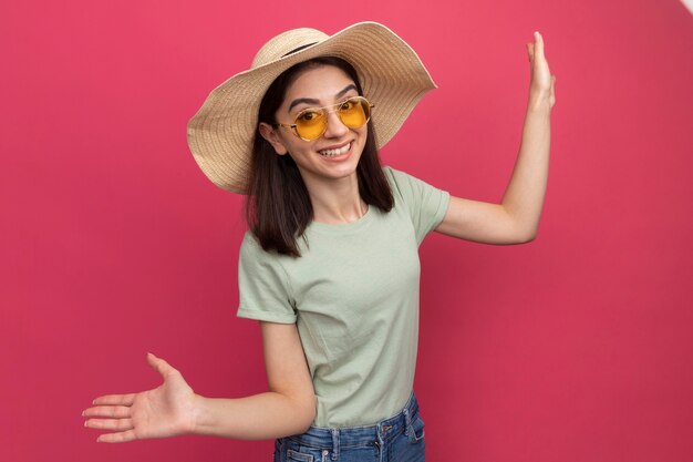 Alegre joven bastante caucásica vestida con sombrero de playa y gafas de sol mostrando las manos vacías