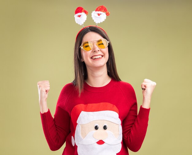 Alegre joven bastante caucásica vistiendo suéter de santa claus y diadema con gafas haciendo gesto de sí con los ojos cerrados aislado sobre fondo verde oliva