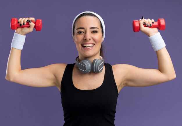 Alegre joven bastante deportiva con diadema y muñequeras con auriculares alrededor del cuello levantando pesas aisladas en la pared púrpura