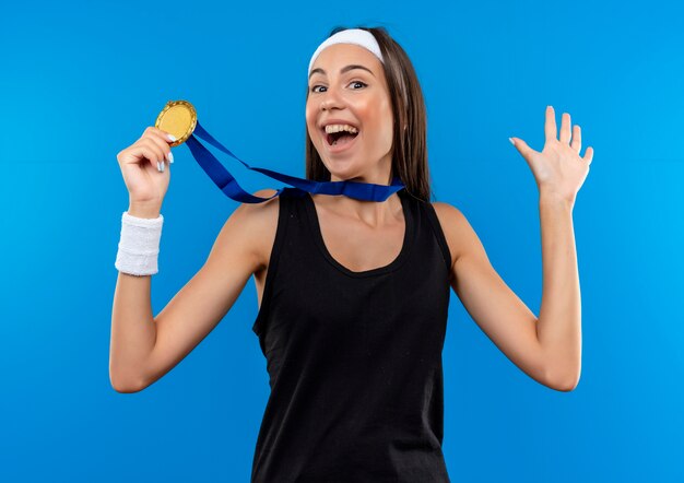 Alegre joven bastante deportiva con diadema y pulsera y medalla alrededor del cuello sosteniendo la medalla y mostrando la mano vacía aislada en el espacio azul