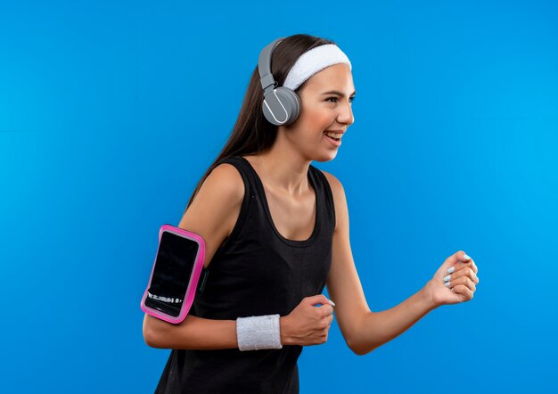 Alegre joven bastante deportivo con diadema y muñequera y auriculares con brazalete de teléfono de pie en la vista de perfil mirando al lado aislado en el espacio azul