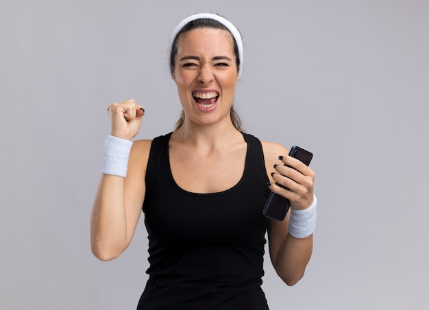 Foto gratuita alegre joven bastante deportivo con diadema y muñequeras sosteniendo el teléfono móvil haciendo sí gesto aislado en la pared blanca con espacio de copia