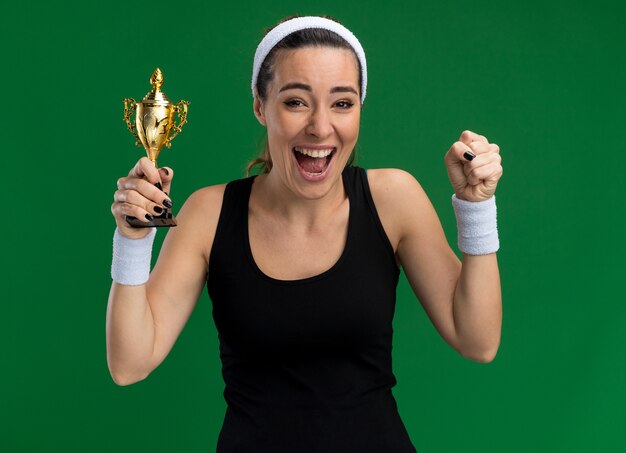 Alegre joven bastante deportivo vistiendo diadema y muñequeras sosteniendo la copa ganadora haciendo gesto de sí