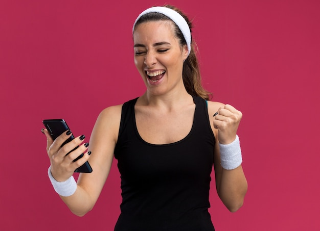 Alegre joven bastante deportivo vistiendo diadema y muñequeras sosteniendo y mirando el teléfono móvil haciendo gesto de sí