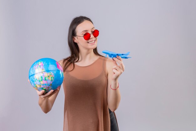 Alegre joven bella mujer con gafas de sol rojas sosteniendo globo y avión de juguete de pie juguetón y feliz