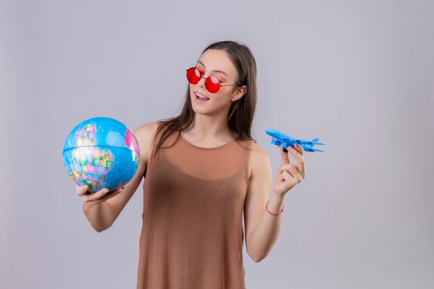 Alegre joven bella mujer con gafas de sol rojas sosteniendo globo y avión de juguete de pie juguetón y feliz