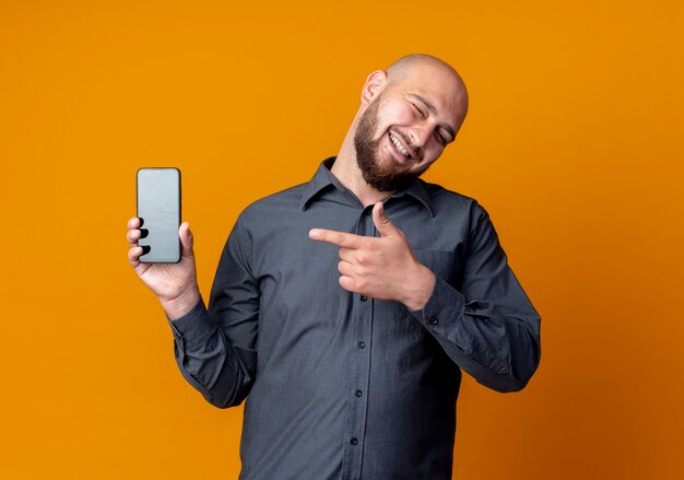Alegre joven calvo call center hombre guiñando un ojo mostrando el teléfono móvil y apuntando a él aislado en naranja